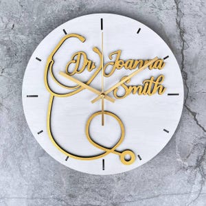 May include: A round white wall clock with gold-colored hands and a gold stethoscope design. The clock face features the text "Dr. Joanna Smith" in a decorative font. Black dashes mark the hours.