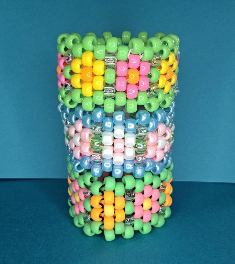May include: A cylindrical kandi cuff bracelet made of colorful plastic beads. The bracelet features a pattern of green, yellow, orange, pink, blue, and white beads. The beads are strung together to form a wearable accessory.