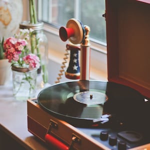 May include: A vintage-style red and white record player with a black vinyl record on the turntable. The record player is open and the lid is propped up. There is a red, white, and blue toy telephone on the table next to the record player.