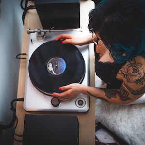 May include: A woman with blue hair and tattoos is playing a black vinyl record on a white turntable. The turntable is on a wooden surface with a red and white striped flag in the background.