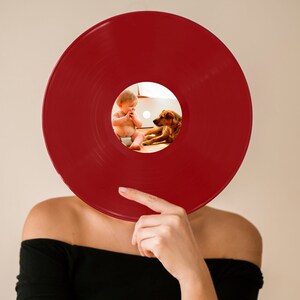 May include: A red vinyl record is held in front of a person's face. A small photo of a baby and a dog is visible through the center hole of the record.