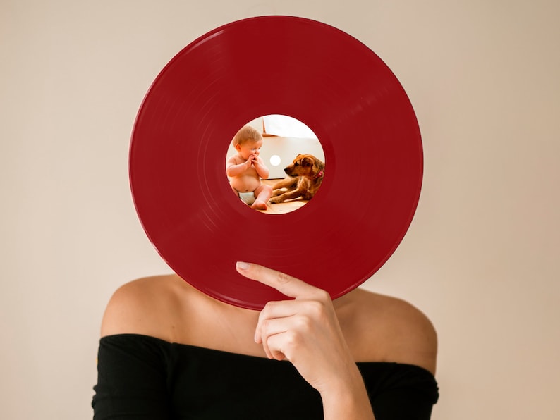 May include: A red vinyl record is held in front of a person's face. A small photo of a baby and a dog is visible through the center hole of the record.