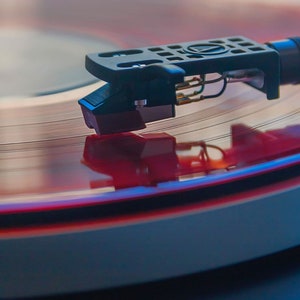 May include: A close-up of a red vinyl record spinning on a turntable. The needle is in the groove, and the record is reflecting the light.