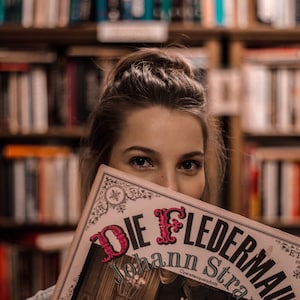 May include: A woman is holding a vintage programme for the opera "Die Fledermaus" by Johann Strauss. The programme is printed on cream-coloured paper with black and red lettering. The background is a blurred image of bookshelves.