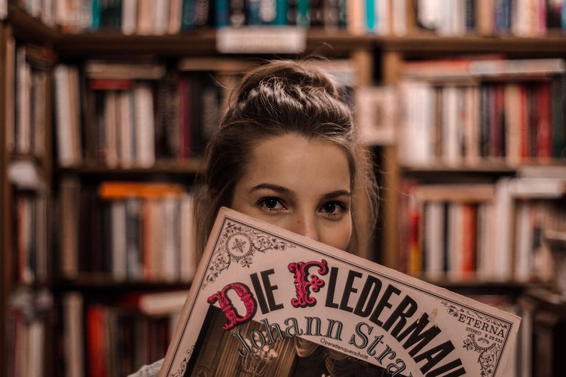 May include: A woman is holding a vintage programme for the opera "Die Fledermaus" by Johann Strauss. The programme is printed on cream-coloured paper with black and red lettering. The background is a blurred image of bookshelves.