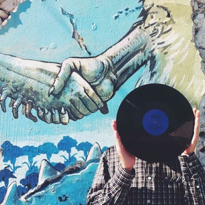 May include: A person is holding a black vinyl record in front of their face. The record has a blue label with the text "Kramo" and "Joy Ride" on it. The person is standing in front of a wall with a colourful mural of two hands shaking.