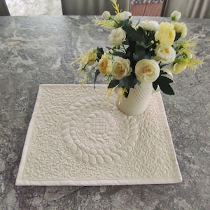 Può includere: Tovaglietta trapuntata color crema con un design circolare centrale e cuciture intricate. Un vaso bianco pieno di rose gialle e bianche è posto sulla tovaglietta. La tovaglietta è quadrata e ha una superficie strutturata.