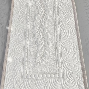 Può includere: Tovaglia da tavolo bianca trapuntata con un motivo di punti decorativi. Il motivo presenta una fila centrale di foglie stilizzate e due file di linee curve su entrambi i lati.