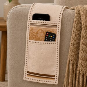 May include: A beige fabric armchair organizer featuring multiple pockets with decorative stitching. The organizer holds a black smartphone and a black remote control. The lower pocket displays a subtle leaf pattern, while the upper section features a tan floral design with brown horizontal stripes.