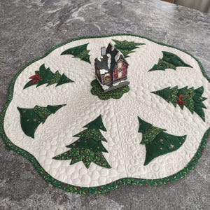 Könnte beinhalten: Eine runde, gesteppte Stoffmatte mit cremefarbenem Hintergrund, wirbelförmigem Steppmuster und gewelltem grünen Rand. Sechs grüne Stoffapplikationen in Form von Weihnachtsbäumen mit goldenen Sternen sind kreisförmig um ein Miniaturhaus angeordnet.