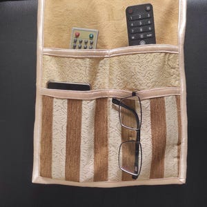 Puede incluir: Un organizador colgante de tela en color beige y marrón con múltiples bolsillos. La sección superior es de color beige sólido, mientras que la inferior presenta rayas verticales marrones y beige. Contiene dos mandos a distancia, un móvil y gafas.