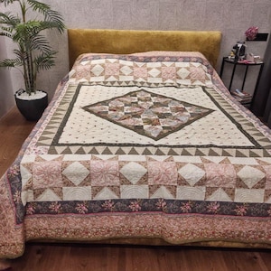 Puede incluir: Una colcha de patchwork con un patrón central de diamantes con motivos florales y geométricos en tonos crema, rosa empolvado y verde oliva. La colcha presenta un borde de triángulos y tela floral, con costuras detalladas en toda la pieza.