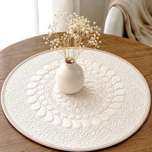 May include: A round, cream-colored placemat with a quilted floral design and a beige border sits on a wooden table. A small, off-white vase holds dried flowers. The table is made of dark wood.