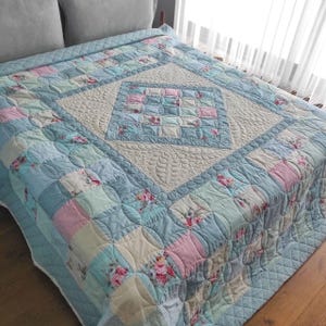 Op de afbeelding: Een patchwork sprei met een centraal ruitvormig paneel met crèmekleurige doorgestikte bladpatronen, omringd door een raster van lichtblauwe, roze en bloemrijke stoffen vierkanten met gebogen randen en gedetailleerde stiksels over het geheel.