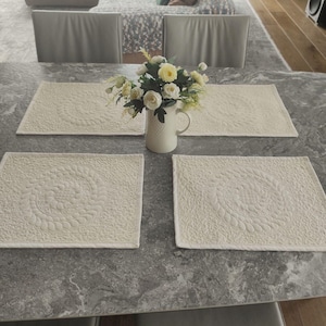 May include: Four white quilted placemats with a circular design on a grey marble table top. A white jug with a floral arrangement of white flowers is in the centre of the table.