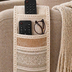 Peut inclure: Organisateur de bras de canapé en tissu beige avec plusieurs poches. Deux télécommandes noires et une paire de lunettes sont dans les poches. L'organisateur a une bordure décorative et est placé sur un accoudoir de canapé beige.