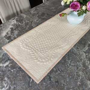 May include: Cream-colored quilted table runner with intricate feather and diamond patterns. The runner has a light tan border and is displayed on a gray countertop. A light blue vase with pink and white flowers sits on the runner.