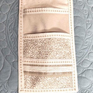 May include: A vertical fabric wall organizer featuring a light beige canvas base with two pockets. The lower pocket displays a tan and white intricate swirling pattern with horizontal textured bands. The edges are finished with decorative stitching.