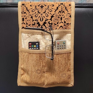 May include: A beige and gold patterned sofa arm organizer with three pockets. The top pocket features a black and gold floral design. The middle pocket holds a pair of glasses and a remote control. The bottom pocket holds a remote control.