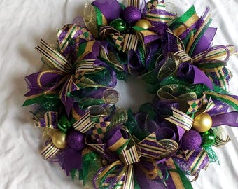 May include: A festive wreath with a circular design, featuring purple, green, and gold ribbons and decorative balls. The wreath is adorned with various textures and patterns, creating a celebratory and eye-catching display.