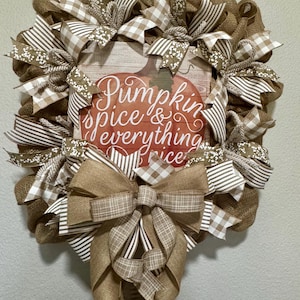 May include: A fall-themed wreath with a burlap base and various ribbons in brown, white, and tan. The wreath features a central square with a pumpkin illustration and the text "Pumpkin spice & everything nice."