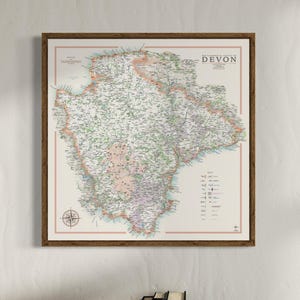 May include: A framed map of the ceremonial county of Devon, England. The map is in a brown frame and has a compass rose in the bottom left corner. The map is titled "The Ceremonial County of Devon" and has a key in the bottom right corner.