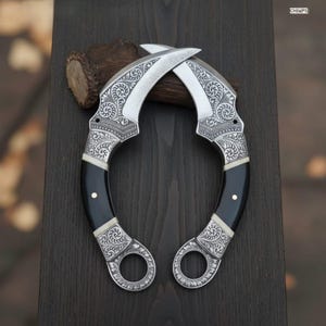 May include: Two curved karambit knives with ornate silver detailing and black handles. The blades are crossed, showcasing the intricate floral patterns etched into the metal. The handles have a white accent and a silver ring at the base.