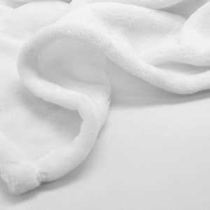 May include: A white plush blanket with a soft, fluffy texture. The blanket is folded and draped over a surface, creating a smooth, flowing curve.