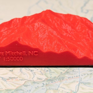 May include: A vibrant red 3D-printed model of Mount Mitchell, NC, with the mountain's name and elevation (6683 feet) inscribed on the front. The model sits on a map, showcasing the topography.