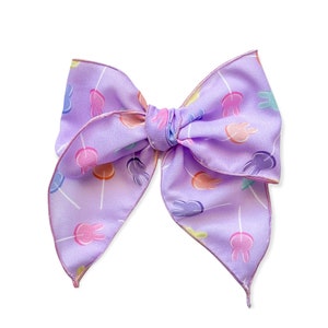 May include: A purple bow with a pattern of colorful bunny lollipops. The bow is made of satin and has a shiny finish.