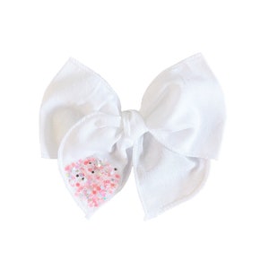 May include: A white fabric bow with a pink and silver glitter heart design.