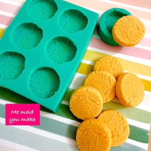 May include: A teal silicone mould with six circular cavities, alongside a smaller green mould and several round, orange objects. The text "Me mold you make" is on a pink rectangle. The objects appear to be handmade.