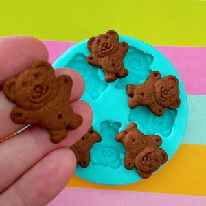 May include: A hand holds a small, brown, bear-shaped biscuit. The biscuit has a smiling face and is next to a turquoise silicone mould with more bear-shaped impressions. The background has colourful stripes.
