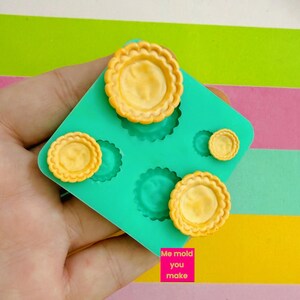 May include: A hand holds a teal silicone mould with several small, yellow, scalloped-edge tart shells. The shells are filled with a lighter yellow material. The text "Me mold you make" is in a pink rectangle.