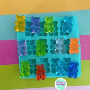 May include: A teal silicone mould filled with colourful gummy bear sweets. The gummy bears are in various colours, including yellow, blue, green, orange, and red. The image also includes a circular sticker with the text "ME MOLD YOU MAKE".