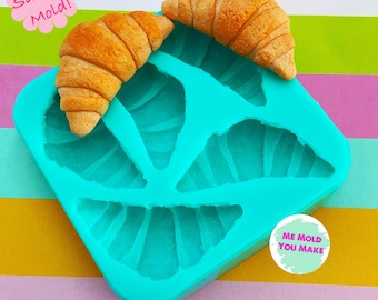 May include: A turquoise silicone mold with three croissant-shaped cavities, accompanied by two miniature, golden-brown croissants. The words "Silicone Mold!" and "ME MOLD YOU MAKE" are also visible.