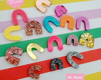 May include: Fifteen pairs of arch-shaped studs in mixed colors and finishes, including glitter, metallic, and solid acrylic. The text on the image reads Arch Studs 15 pairs of Mixed colours and shapes and Me Mold You Make. These are small craft components.