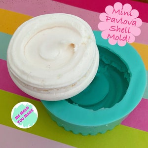 May include: A white mini pavlova shell beside a teal silicone mould. The mould has a scalloped edge and the text "Mini Pavlova Shell Mold!" in pink. A round sticker with the text "ME MOLD YOU MAKE" is also visible.