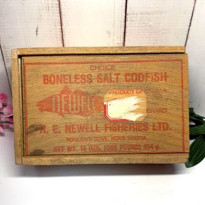 Könnte beinhalten: Eine rechteckige Holzkiste mit rotem Aufdruck, der Choice Boneless Salt Codfish, Product of Canada, Newell Brand, R. E. Newell Fisheries Ltd., Vogler's Cove, Nova Scotia und Net Wt. 16 ozs. (One Pound) 454 g. lautet.
