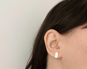 May include: A small, rectangular silver stud earring with a smooth, polished finish. The earring features a flat, vertical bar design that sits securely against the earlobe, providing a clean and simple aesthetic for everyday wear.