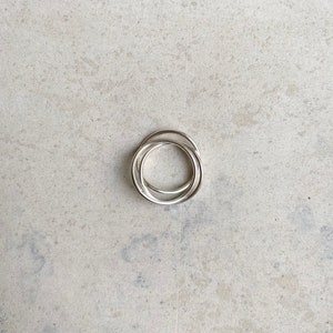 Könnte beinhalten: Ein silberfarbener Ring, der aus drei ineinandergreifenden, dünnen Bändern besteht. Das Metall hat eine glatte, polierte Oberfläche und eine kreisförmige Form. Die drei einzelnen Bänder sind zu einem zusammenhängenden Schmuckstück verbunden.