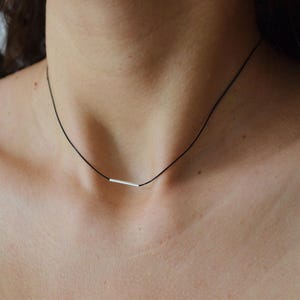 May include: A thin black cord necklace featuring a single, small, horizontal silver-toned metal bar pendant centered on the cord. The necklace rests against the neck, showcasing the contrast between the dark cord and the metallic bar element.