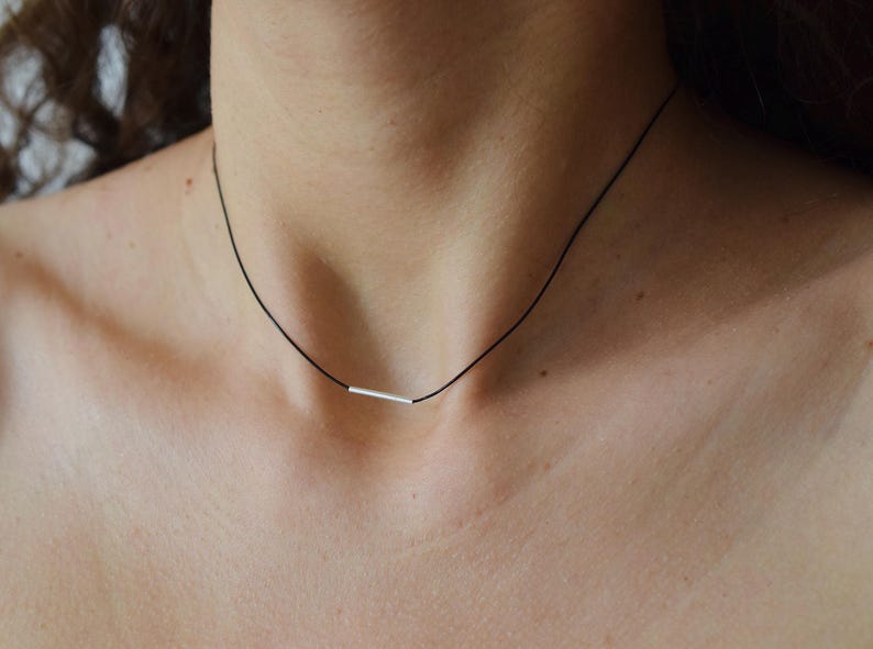 May include: A thin black cord necklace featuring a single, small, horizontal silver-toned metal bar pendant centered on the cord. The necklace rests against the neck, showcasing the contrast between the dark cord and the metallic bar element.