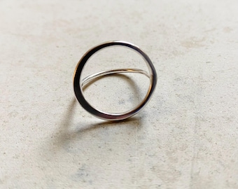 May include: A silver ring featuring a circular design with a smooth, polished finish. The band is thin and forms a continuous open circle shape, reflecting light on its metallic surface. The ring is captured from a slightly elevated angle.