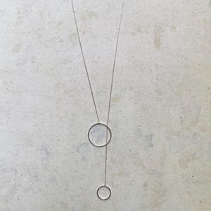 Puede incluir: Un collar plateado con dos colgantes circulares. El círculo más grande está en la parte superior, con un círculo más pequeño suspendido debajo. El collar cuelga sobre un fondo claro y texturizado.