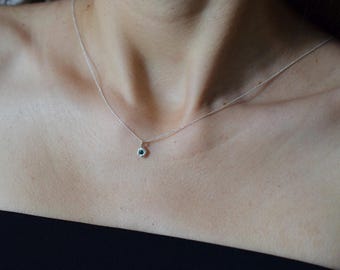 May include: A delicate silver chain necklace featuring a small, round, teal-colored gemstone pendant set in a simple metal bezel. The necklace rests against the collarbone, showcasing a thin, polished silver link chain and a single, centered stone.