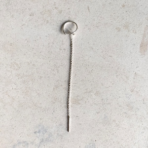 Puede incluir: Un pendiente de cadena de plata con un pequeño aro en la parte superior y una cadena larga y fina que termina en una pequeña barra recta.