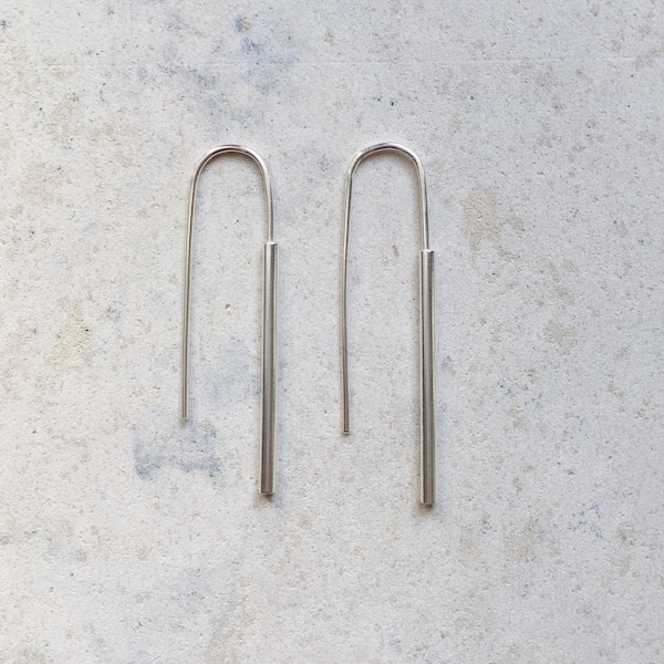 Puede incluir: Un par de pendientes de tono plateado con un diseño de alambre en forma de U y una barra cilíndrica lisa unida a un extremo. Los pendientes tienen un acabado metálico pulido y una estructura lineal limpia que cuelga verticalmente.
