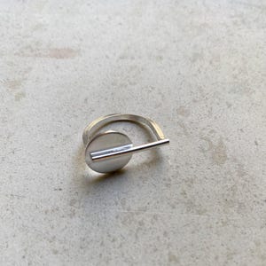 Könnte beinhalten: Ein silberfarbener Ring mit einem kreisförmigen Scheibenelement, das an einem dünnen, horizontalen Stab befestigt ist, der über die Vorderseite verläuft. Das Band ist glatt und metallisch und weist eine polierte Oberfläche auf.