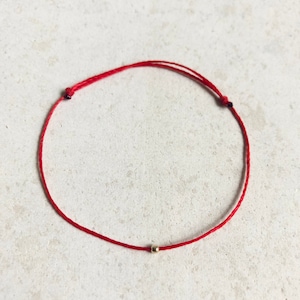 Può includere: Un braccialetto di corda rossa con un piccolo perno dorato.